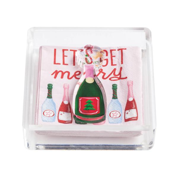 Clear acrylic napkin holder with 'Let's Get Merry' napkins and a champagne bottle weight.