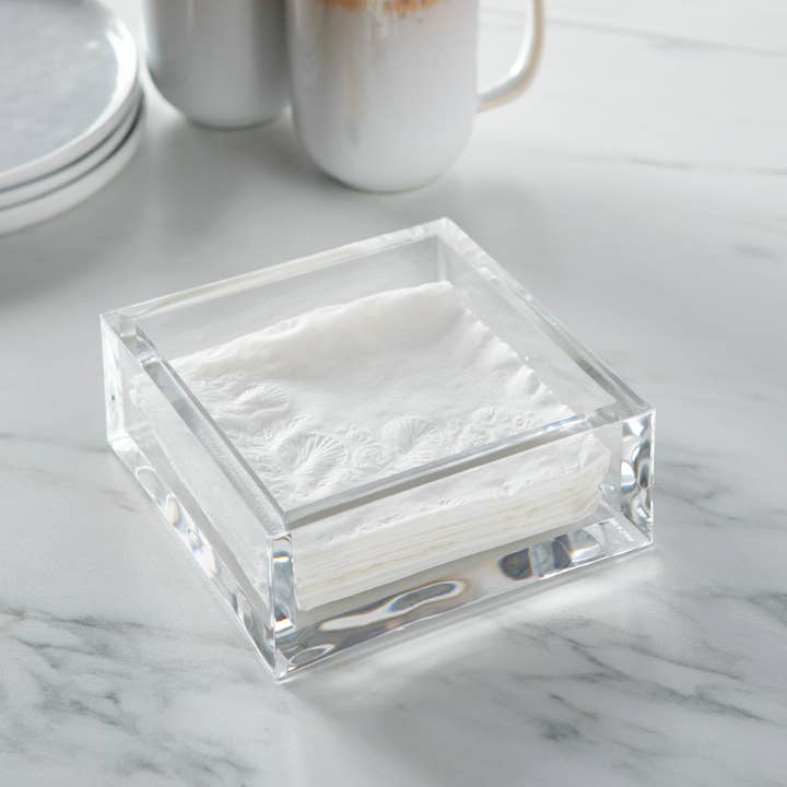 Clear acrylic square cocktail napkin holder with white napkins on a marble surface.