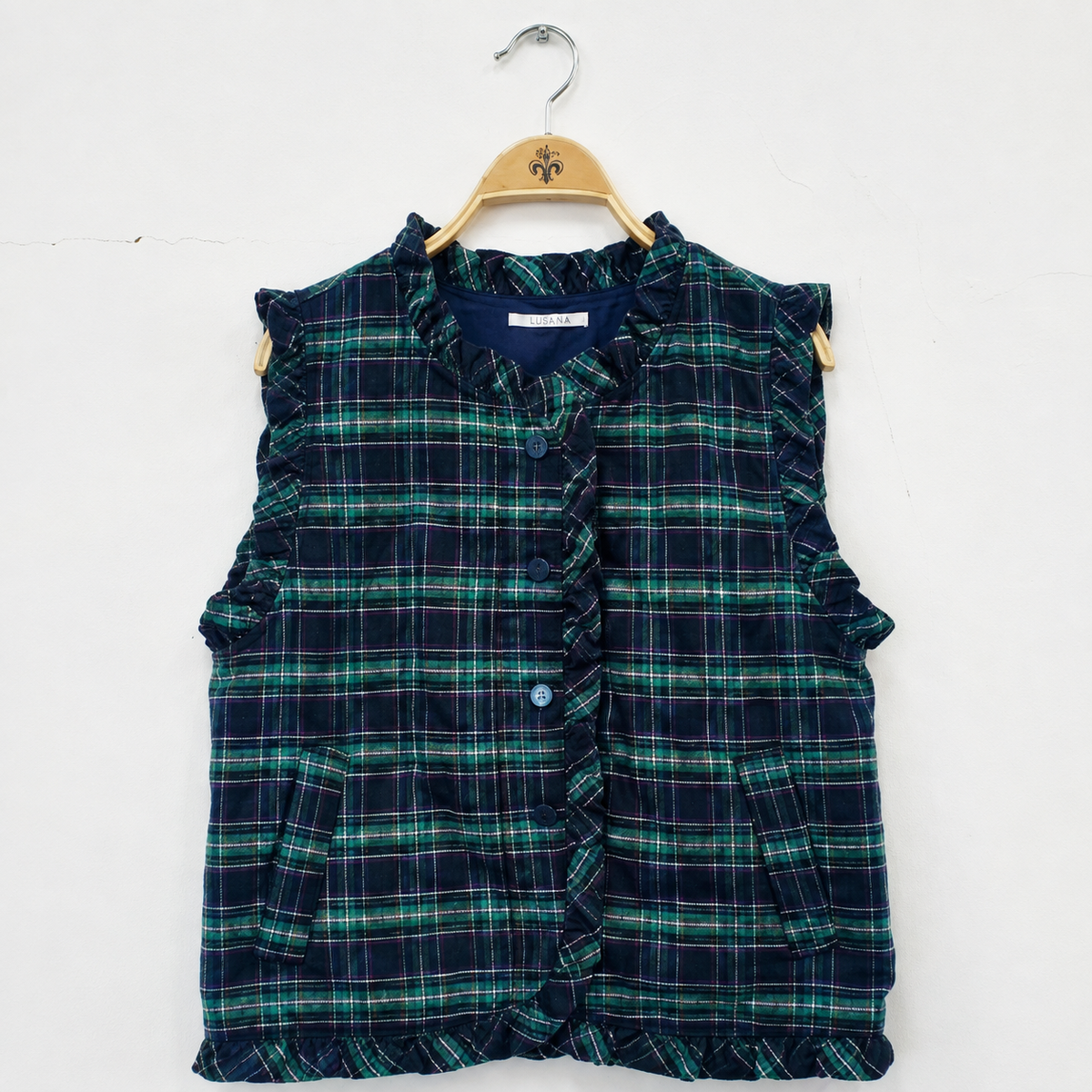 Green and navy plaid quilted vest with ruffle trim, button front, and side pockets on a hanger.
