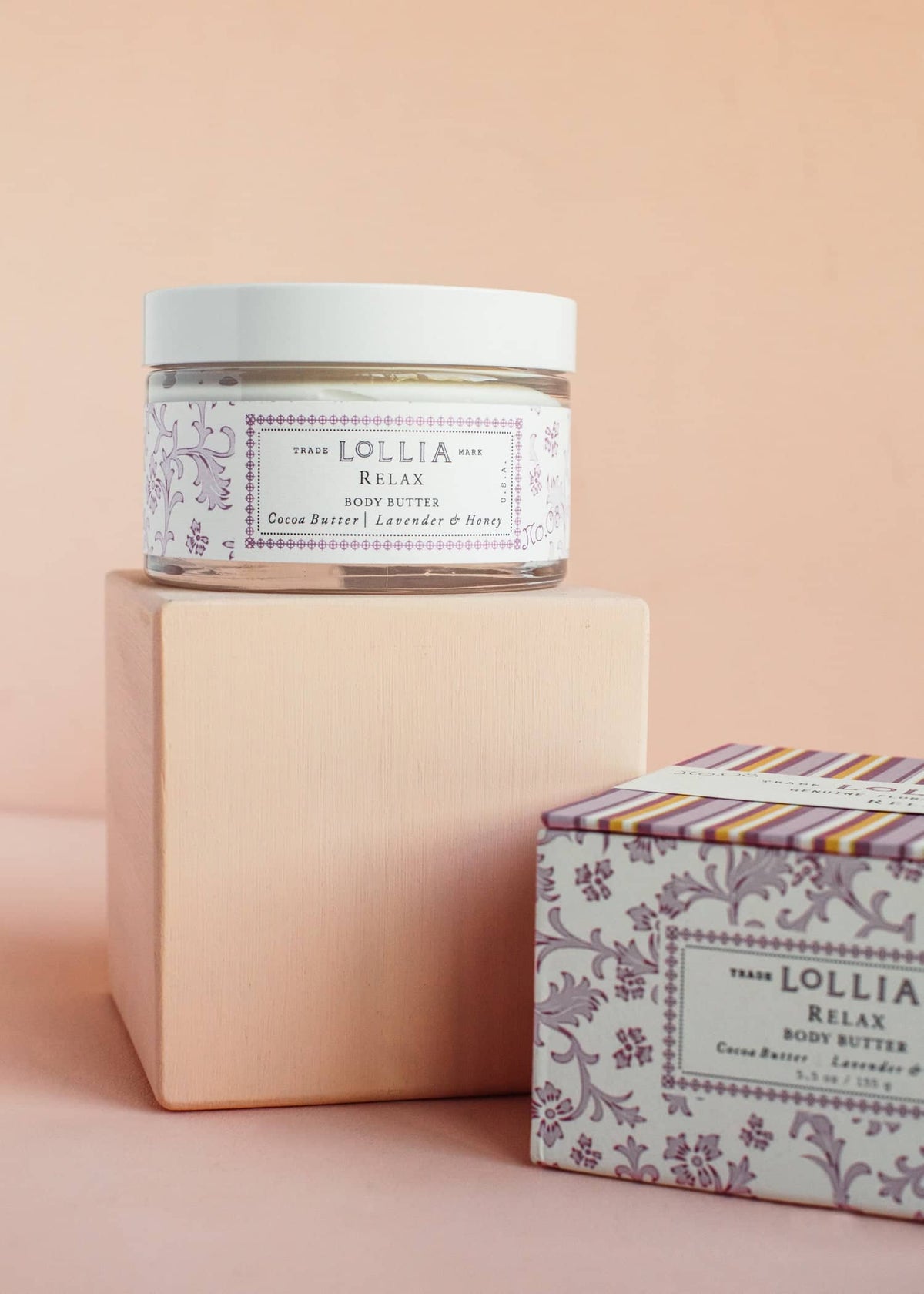 Lollia Relax Body Butter jar on a wooden block next to its floral and striped packaging box.