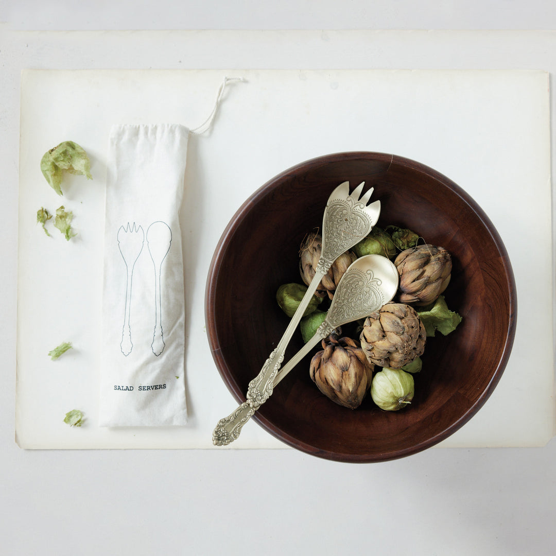 Embossed brass salad servers in a wooden bowl next to a printed drawstring storage bag.