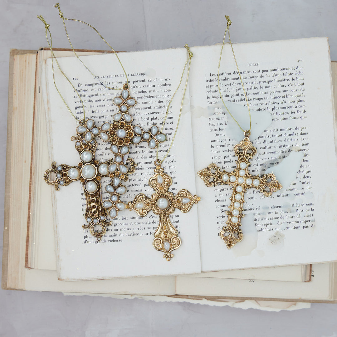 Four ornate gilded pewter cross ornaments with faux pearl accents resting on an open vintage book.