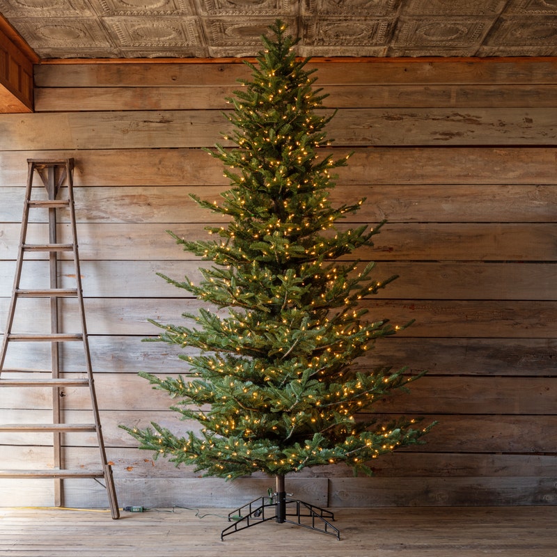 10-foot Grand Noble artificial Christmas tree with warm lights and a metal stand in a rustic room.