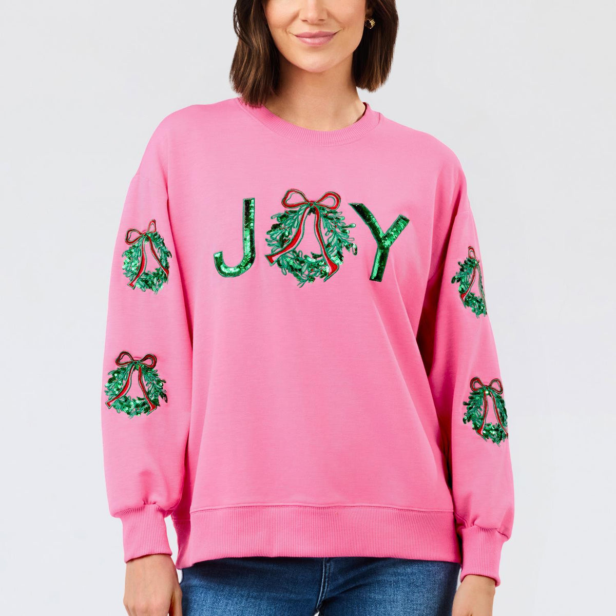 Woman wearing a pink Sarah Joy Wreath sweatshirt with green sequin 'JOY' and wreath designs.