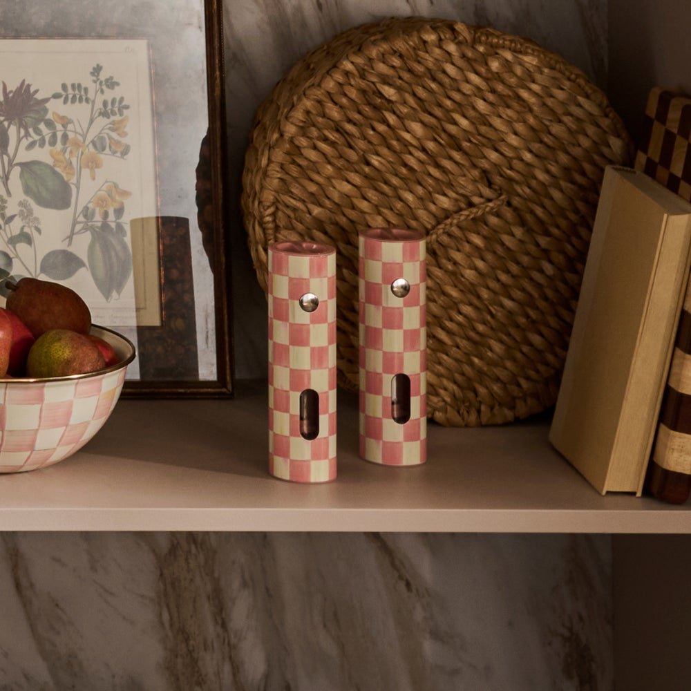 Two Rosy Check electric salt and pepper grinders on a shelf next to a bowl of fruit and a book.