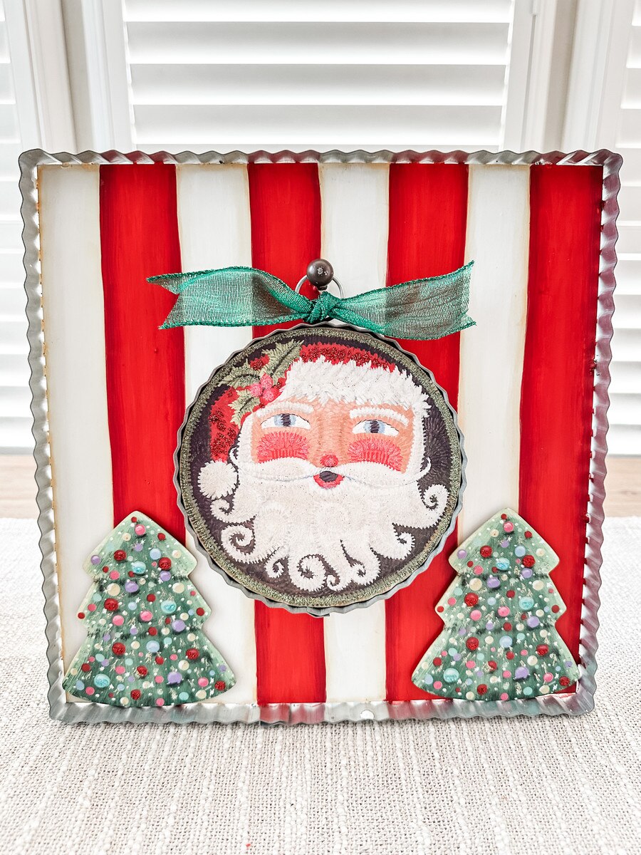 Square red and white striped metal display board with a Santa ornament and two small green trees.