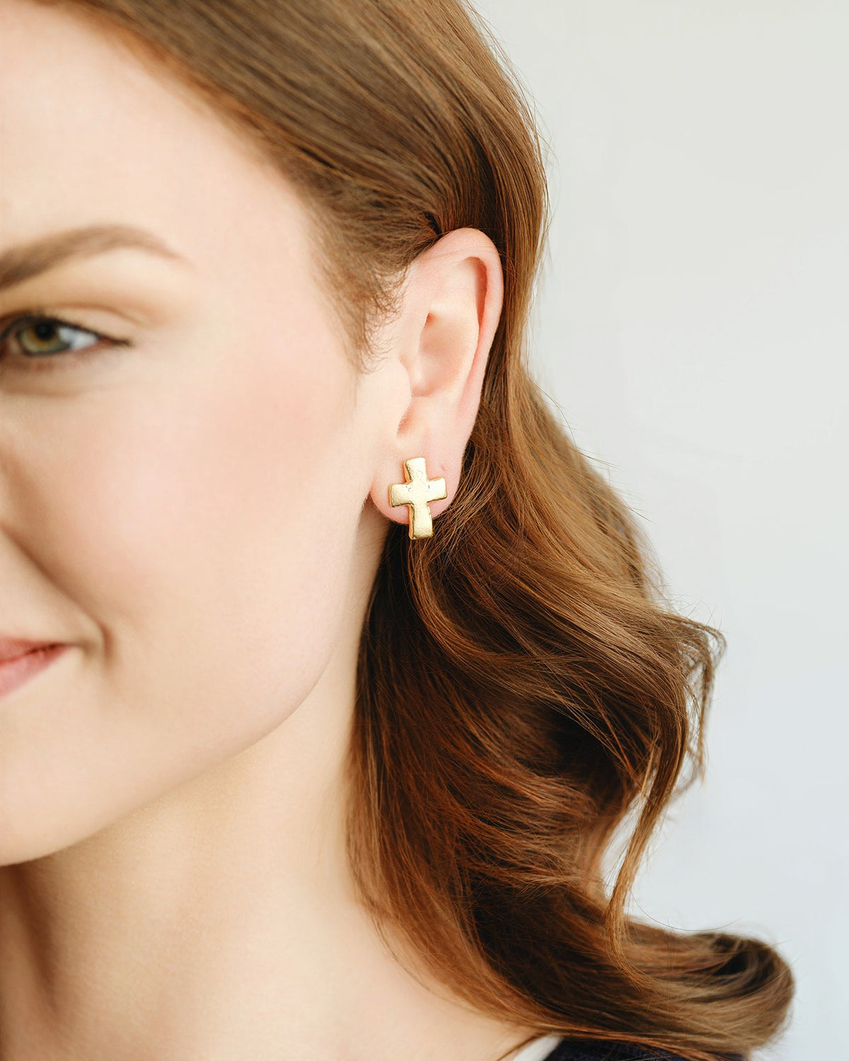 A person wearing a small, gold-plated cross stud earring on their earlobe.