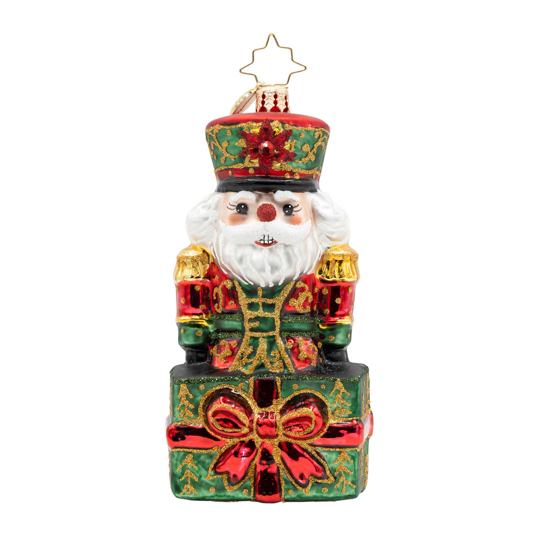 The Gifted Guard nutcracker glass ornament popping out of a green gift box with a red bow.