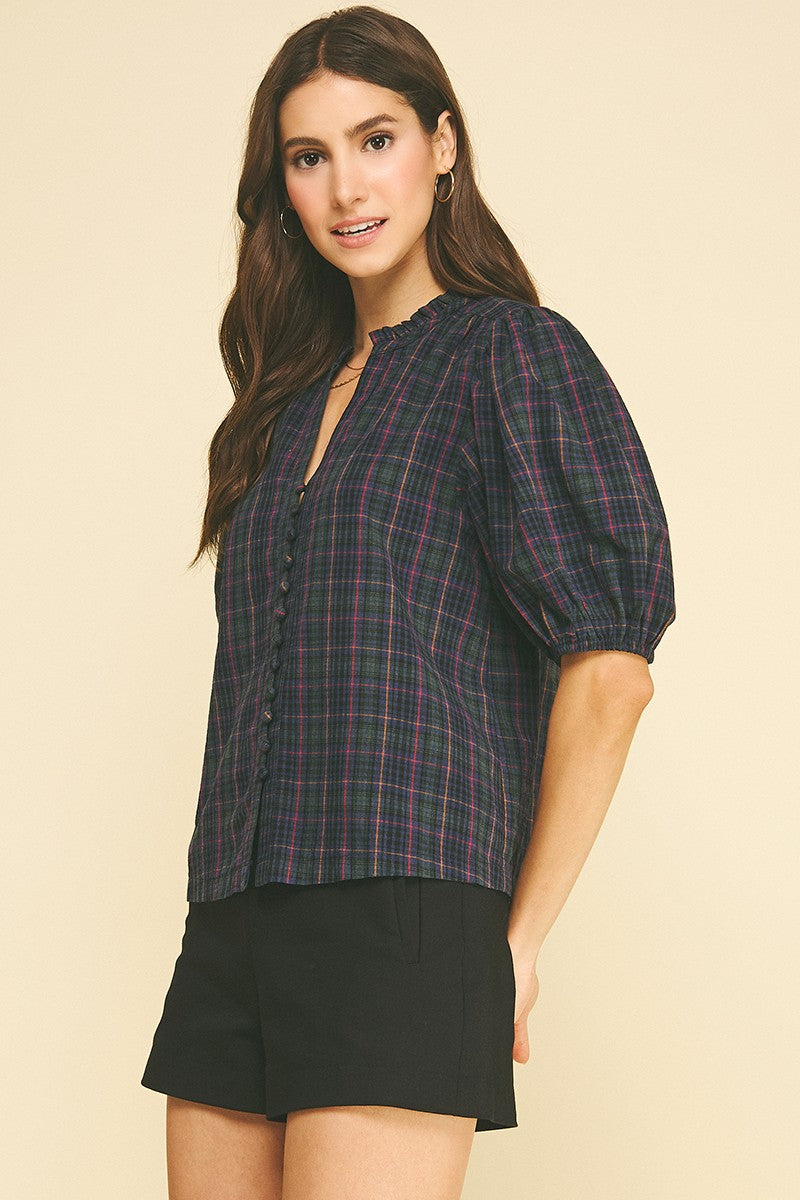 Woman wearing the April Plaid Puff Sleeve Blouse in a dark navy and green plaid with black shorts.