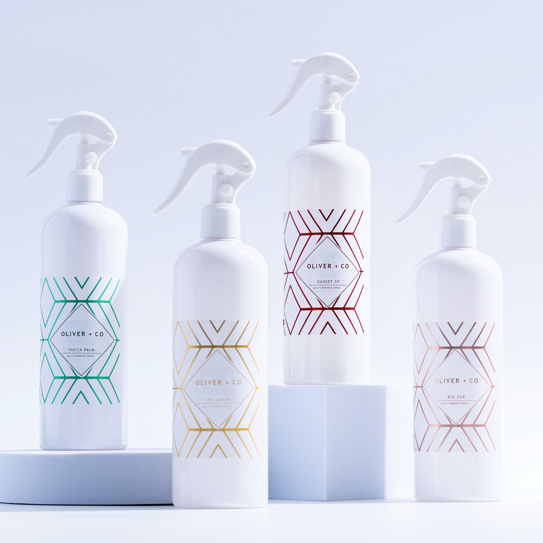 Four white Oliver + Co Multi-Purpose Cleaner spray bottles with geometric designs in various colors.