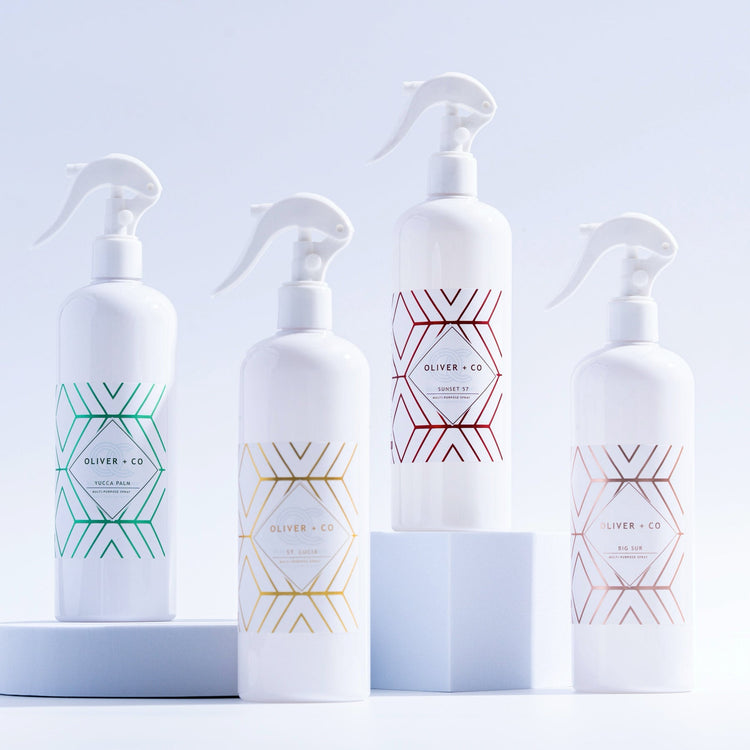 Four white Oliver + Co Multi-Purpose Cleaner spray bottles with geometric designs in various colors.