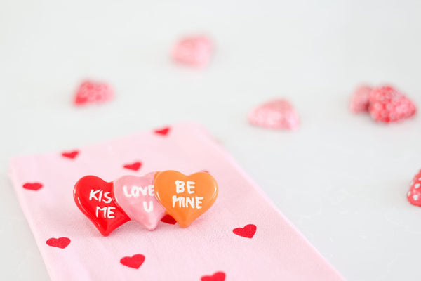 Three heart-shaped charms with 'KISS ME', 'LOVE U', and 'BE MINE' on a pink heart-patterned cloth.