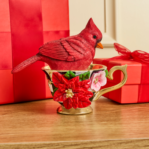 A red cardinal figurine sitting inside a decorative floral teacup on a wooden surface.