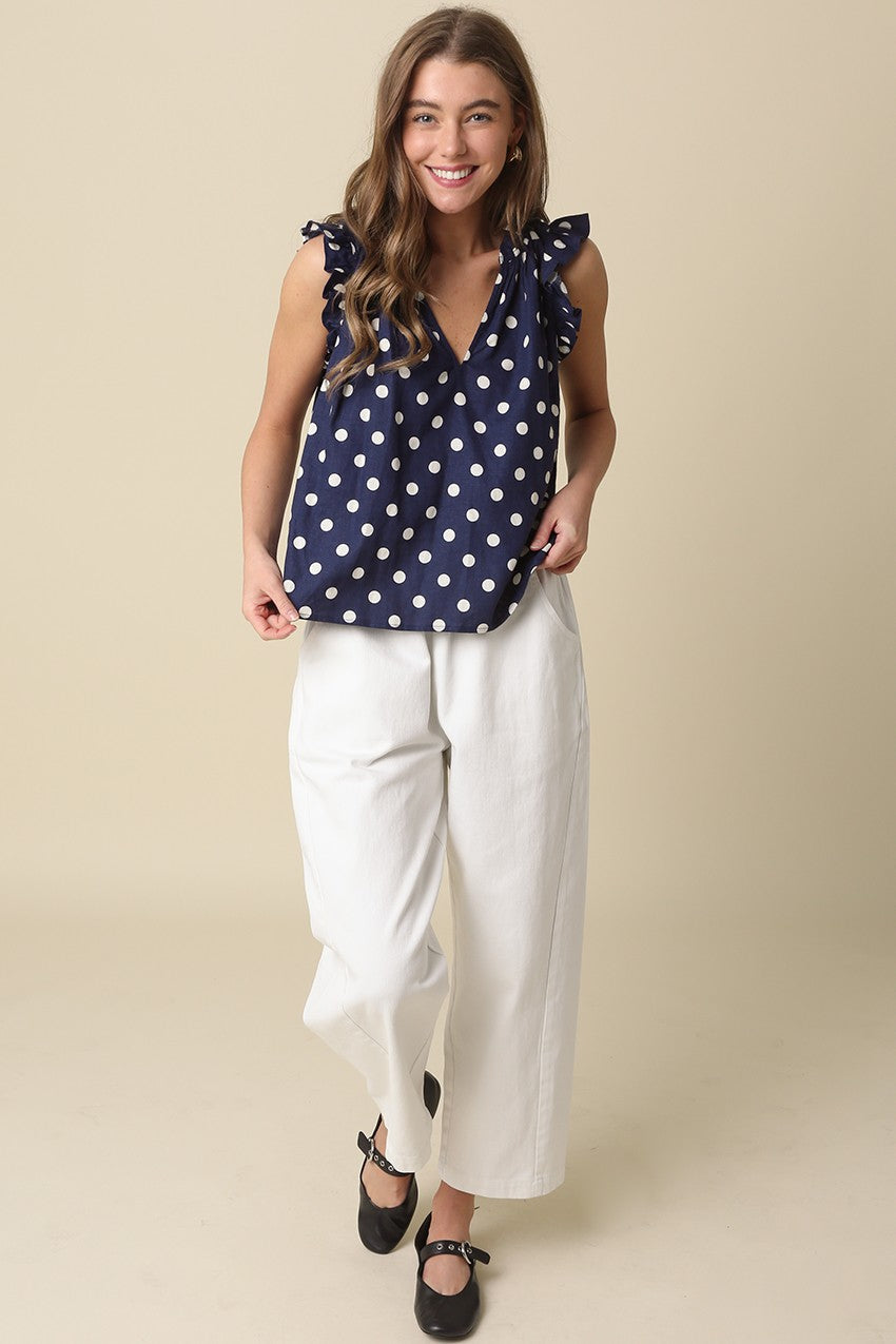 Woman wearing the Mira navy blue polka dot ruffle blouse with white wide-leg pants.