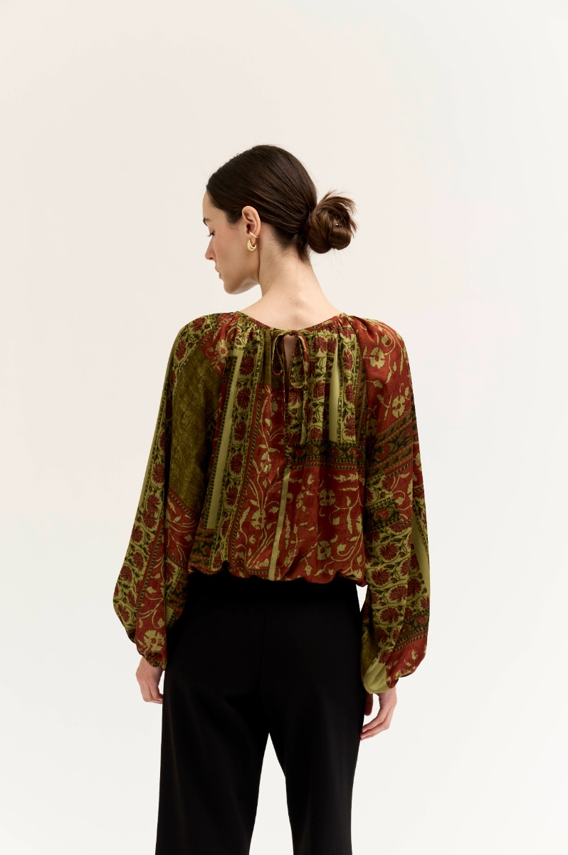 Back view of a woman wearing the Amy Printed Voluminous Top in olive and rust with black trousers.